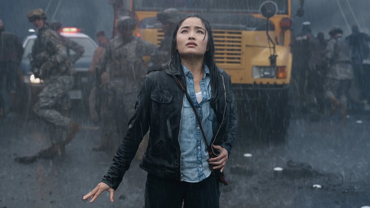 Cate (Anna Sawai) stands on a rainy bridge looking up at Godzilla with a school bus and soldiers behind her.